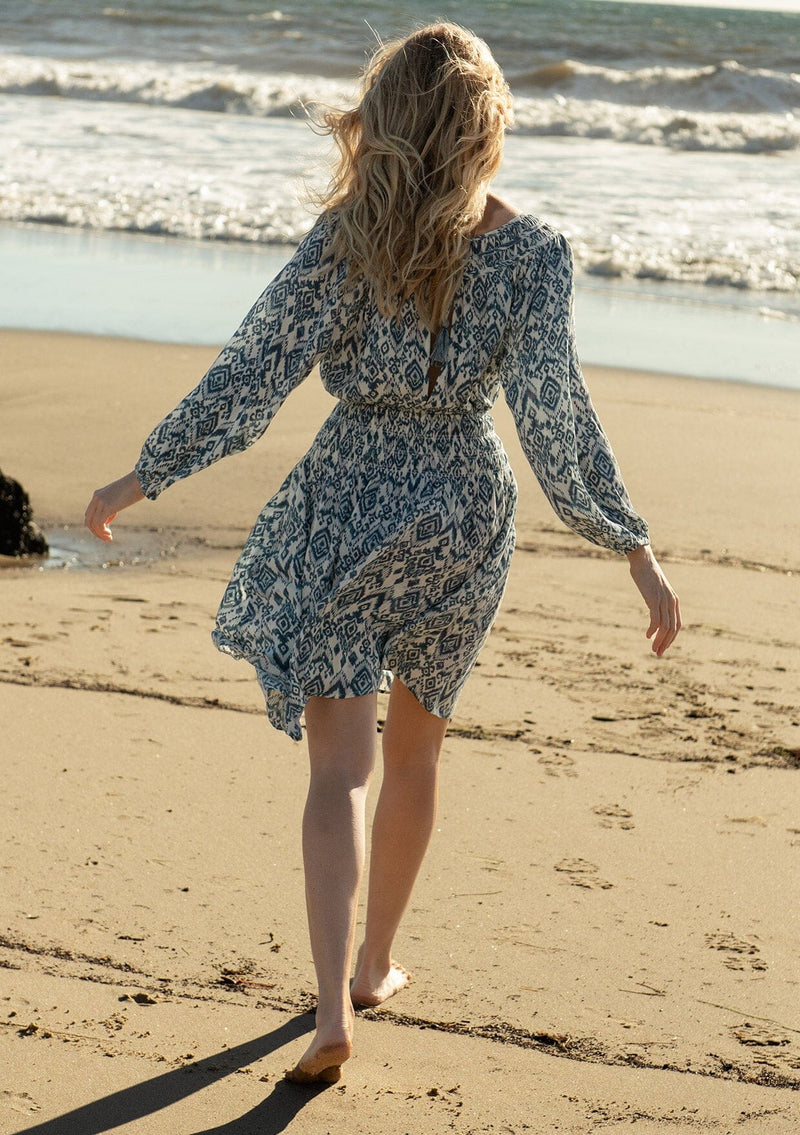 [Color: Ivory/Denim] A back facing image of a blonde model at the beach wearing a flowy spring mini dress in a bohemian blue and white abstract diamond print. With voluminous long sleeves, a smocked elastic waist, an open back with tassel tie closure, and a v neckline detail. 
