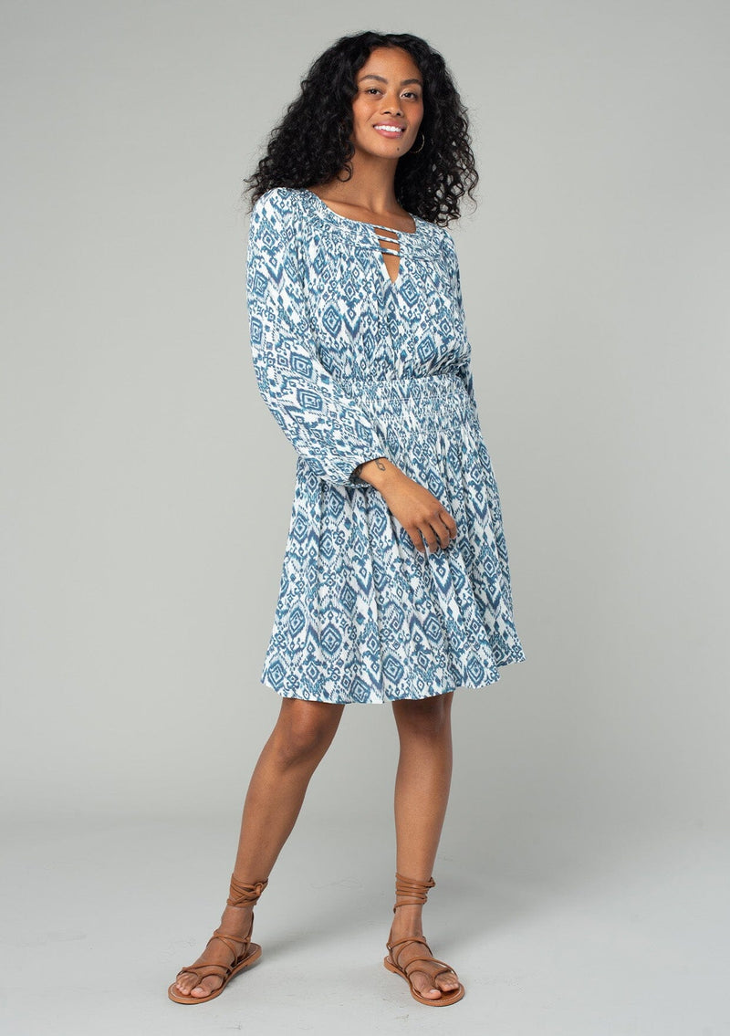 [Color: Ivory/Denim] A full body front facing image of a brunette model wearing a flowy spring mini dress in a bohemian blue and white abstract diamond print. With voluminous long sleeves, a smocked elastic waist, an open back with tassel tie closure, and a v neckline detail. 