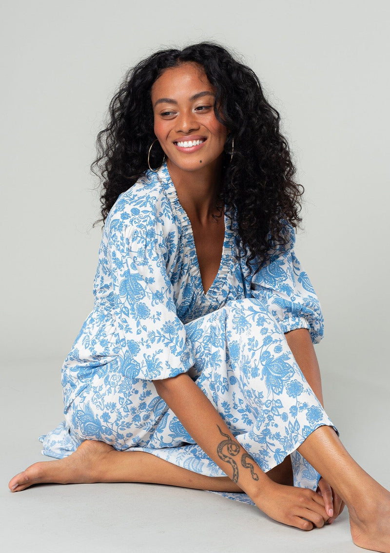 [Color: Cream/Dusty Blue] A sitting front facing image of a brunette model wearing a bohemian cottage core style maxi dress in a white and blue floral print. With half length puff sleeves, a smocked elastic waist detail, a flowy tiered skirt, and a v neckline. 
