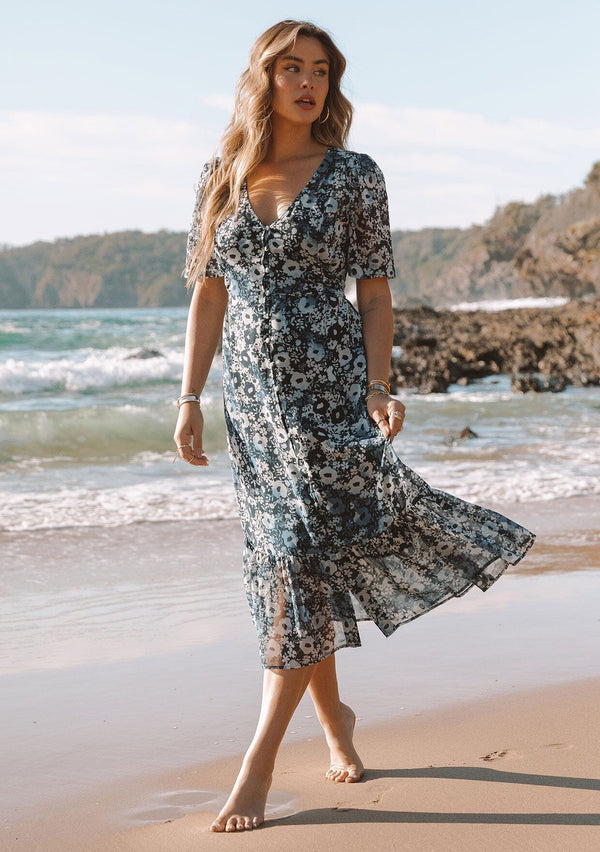 [Color: Indigo/Blue] A full body front facing image of a blonde model wearing a pretty bohemian mid length chiffon dress in a blue and navy blue floral print. With short sleeves, a self covered button front, a v neckline, and a back waist tie.