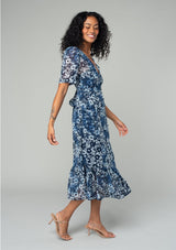 [Color: Indigo/Blue] A side facing image of a brunette model wearing a pretty bohemian mid length chiffon dress in a blue and navy blue floral print. With short sleeves, a self covered button front, a v neckline, and a back waist tie. 