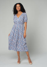 [Color: Blue/Coral] A front facing image of a brunette model wearing a bohemian spring mid length dress in a blue and coral floral print. With short puff sleeves, a button front, and side pockets. 