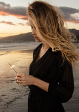 [Color: Black] A close up side facing image of a blonde model on the beach wearing a bohemian holiday mini dress in a black lurex stripe. With long sleeves, a tiered flowy skirt, a smocked elastic waist, a v neckline, and an open back with tie closure.