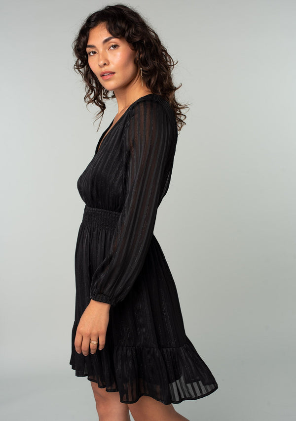 [Color: Black] A side facing image of a brunette model wearing a bohemian holiday mini dress in a black lurex stripe. With long sleeves, a tiered flowy skirt, a smocked elastic waist, a v neckline, and an open back with tie closure.