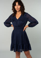 [Color: Navy] A front facing image of a brunette model wearing a bohemian holiday mini dress in a navy blue lurex stripe. With long sleeves, a tiered flowy skirt, a smocked elastic waist, a v neckline, and an open back with tie closure. 