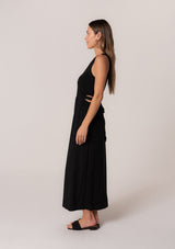 [Color: Black] A side facing image of a brunette model wearing a soft and stretchy knit sleeveless maxi dress in black. With a v neckline, sexy side waist cutouts, and a self tie belt. 