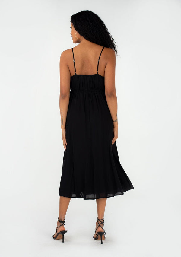 [Color: Black] A back facing image of a brunette model wearing a sexy spring slip dress in black. With adjustable spaghetti straps, a v neckline, lace trim, a half elastic waist at the back, and a flowy paneled mid length skirt. 