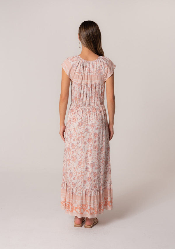 [Color: Natural/Clay] A back facing image of a brunette model wearing a pink floral bohemian maxi dress. With short cap sleeves, an elastic waist, a tiered flowy skirt, a v neckline, and double tassel tie neckline. 