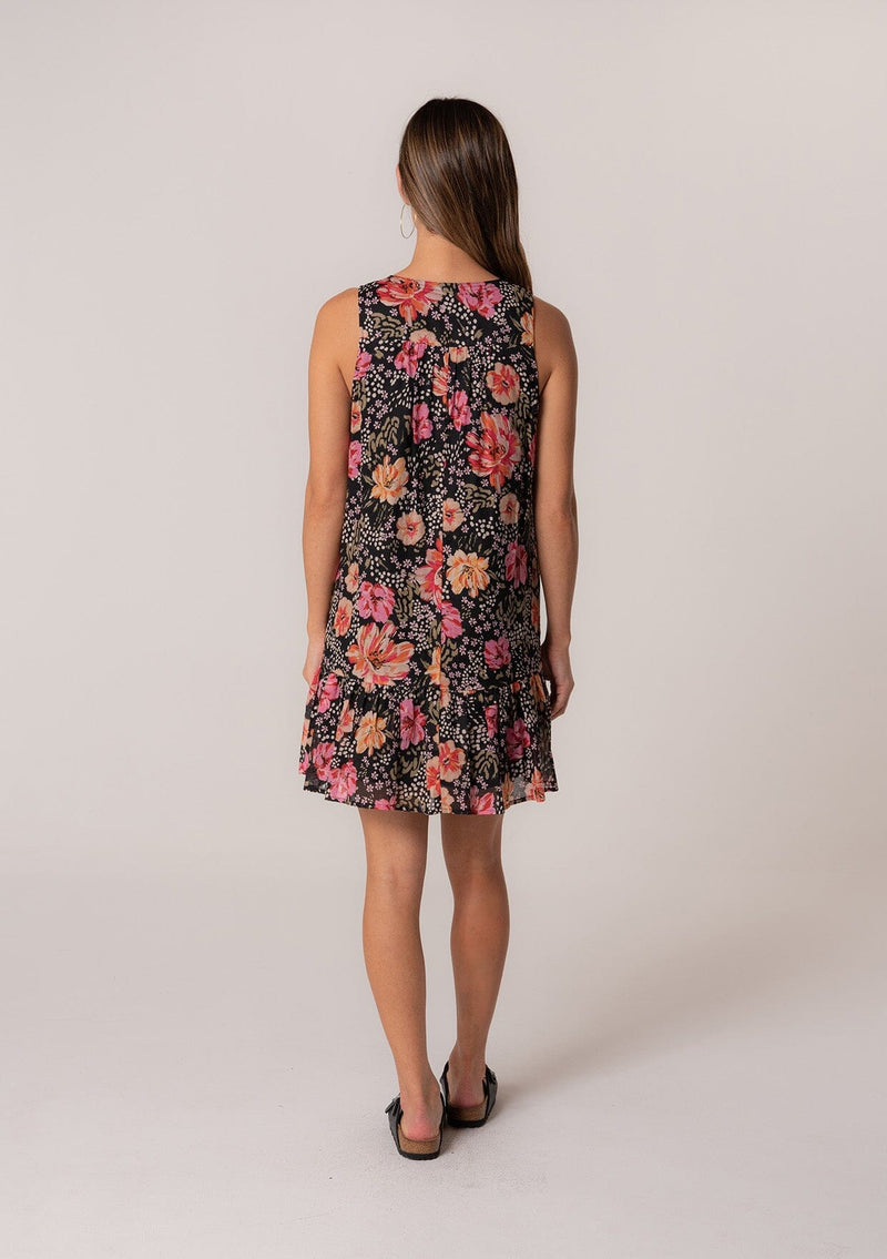 [Color: Black/Fuchsia] A back facing image of a brunette model wearing a summer bohemian cotton mini dress in a black and pink floral print. A sleeveless mini dress with a roomy silhouette, a v neckline, and a tiered skirt. 