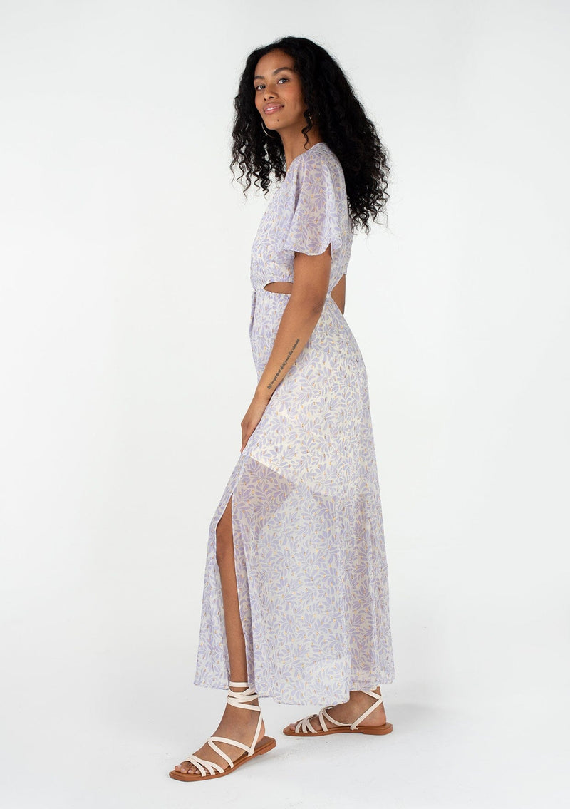 [Color: Natural/Lilac] A side facing image of a brunette model wearing a sheer chiffon bohemian maxi dress in an off white and light purple leaf print. With gold clip dot details, short flutter sleeves, a v neckline, side waist cut outs, and a back keyhole. 