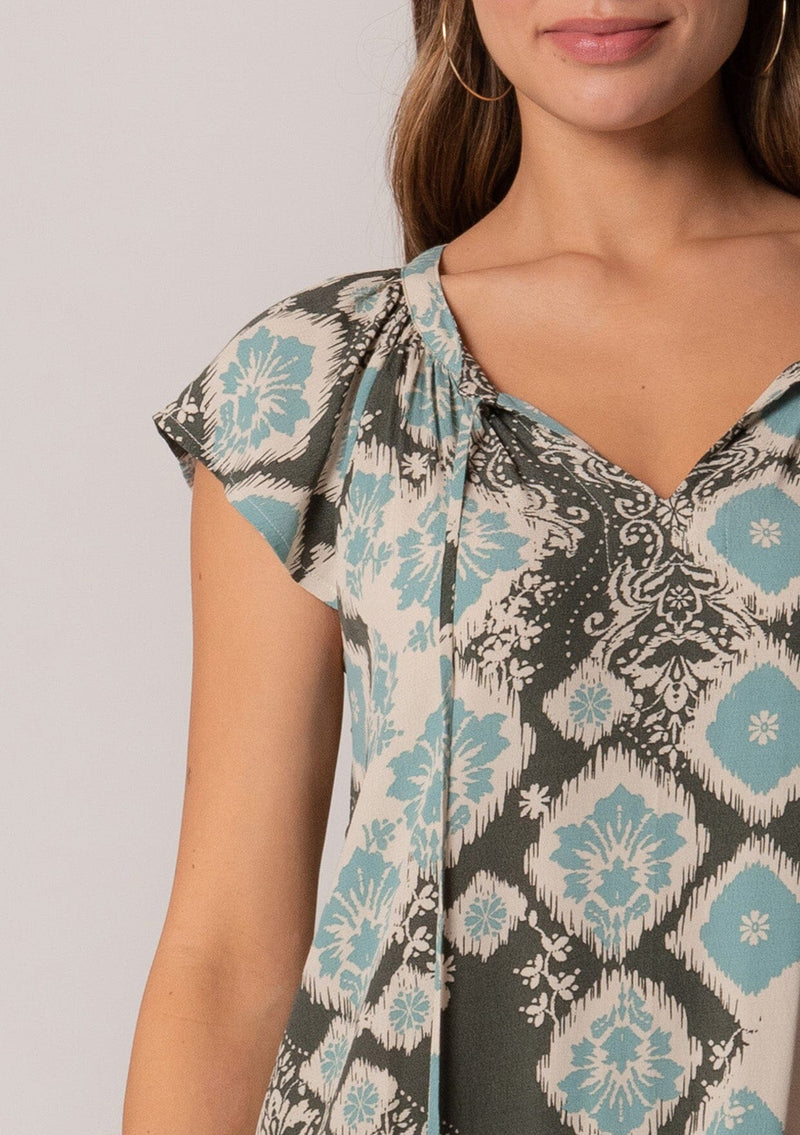 [Color: Black/Aqua] A close up front facing image of a brunette model wearing a lightweight summer mini dress in a green bohemian print. With short flutter sleeves, a split v-neckline with ties, and a ruffle trimmed tiered skirt. 