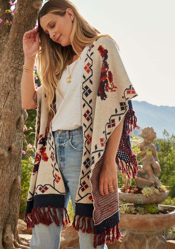 [Color: Floral] A model wearing a red and blue floral sweater poncho with fringed detail and a contrast open knit back. 