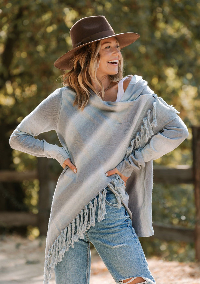 [Color: Chambray/Offwhite] A model wearing a light blue and off white space dye stripe sweater. With a side button closure, long sleeves, and a fringed waterfall hemline. 