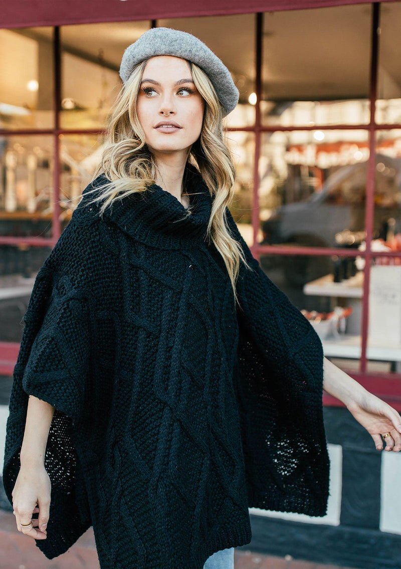 [Color: Black] A blond woman outside wearing an ultra soft chunky cable knit poncho sweater. Featuring a cozy cowl neckline.