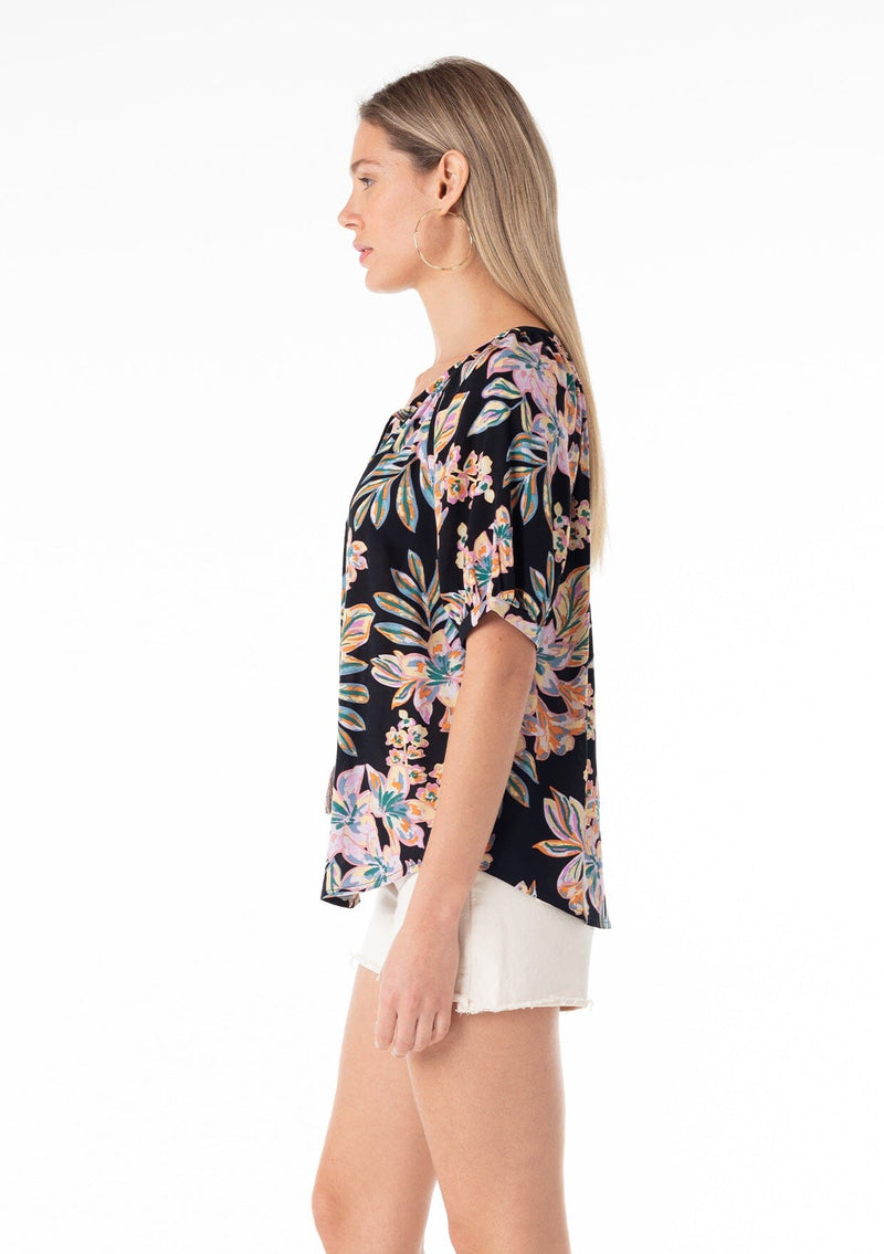 [Color: Black/Pink] A side facing image of a blonde model wearing a best selling short puff sleeve blouse in a black and pink floral print. With a self covered button front, a split v neckline, tassel ties, and a relaxed fit. 