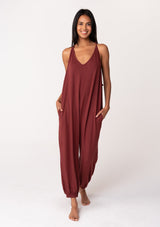 [Color: Rust] A front facing image of a brunette model wearing a cozy sleeveless, French terry jumpsuit with tie back detail, side pockets, and deep v neckline. A lounge jumpsuit in dark red. 