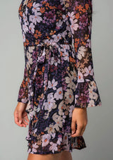 [Color: Black/Dusty Rose] A front facing image of a brunette model wearing a chiffon bohemian mini wrap dress in a black and purple floral print. With long bell sleeves, a ruffled hemline, and a side tie closure.