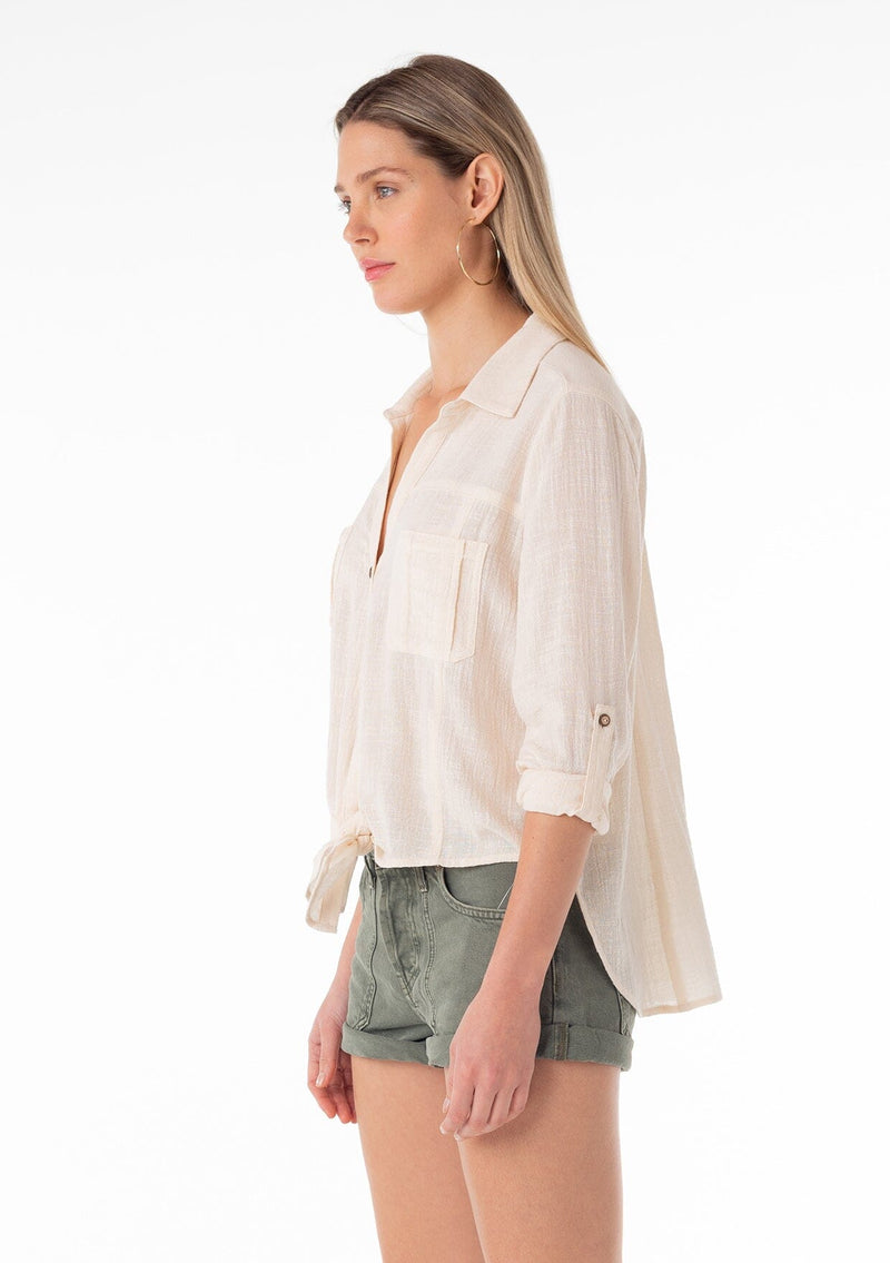[Color: Natural] A side facing image of a blonde model wearing a natural, off white cotton shirt. With long rolled sleeved, a button tab sleeve closure, a button front, two front patch pockets, a tie front waist detail, and a high low hemline.