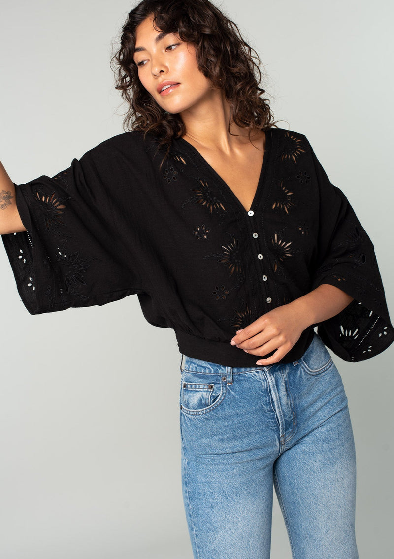 [Color: Black] A half body front facing image of a brunette model wearing a bohemian black embroidered eyelet top with billowy half length sleeves, a button front, and a v neckline. 