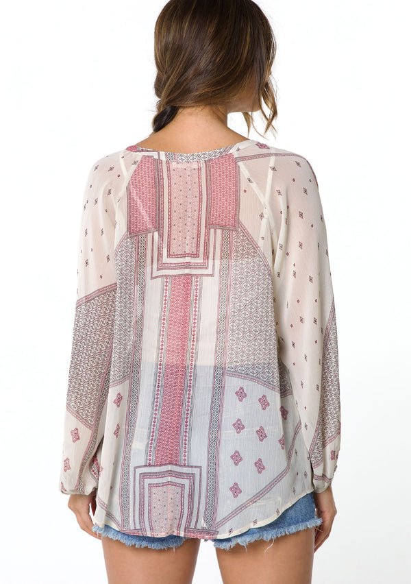 [Color: Natural/Wine] A model wearing a sheer chiffon peasant top in a pink and natural mixed bohemian print. With long voluminous sleeves, a split neckline with tassel ties, and a button front.