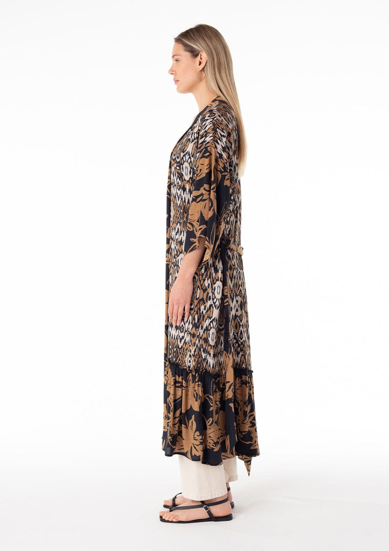 [Color: Black/Taupe] A side facing image of a blonde model wearing a mid length duster lounge robe in a black and taupe mixed floral and ikat print. With half length long sleeves, a ruffle trimmed tiered hemline, an open front, and a tie waist belt.