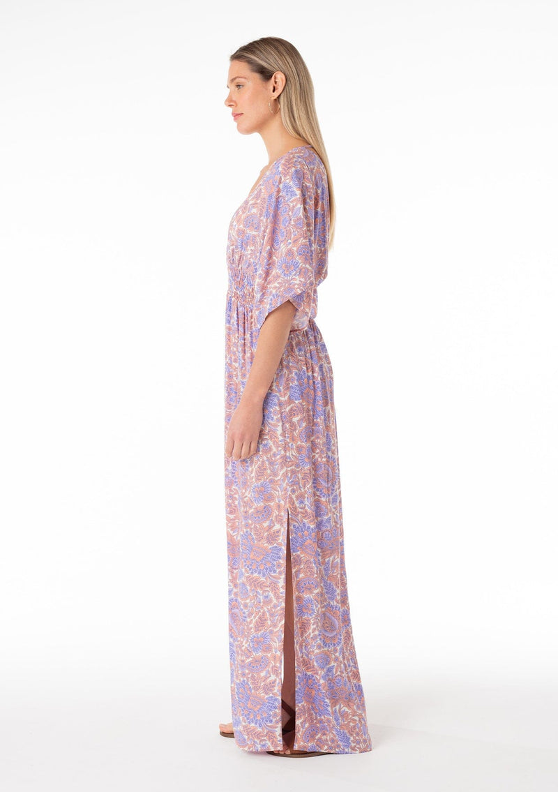[Color: Ivory/Coral] A side facing image of a blonde model wearing a best selling resort bohemian maxi dress in a purple and coral pink retro floral print. With short kimono sleeves, a surplice v neckline, a smocked elastic waist, and an open back with tie closure. 