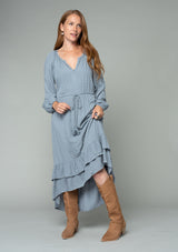 [Color: Grey] A front facing image of a red headed model wearing a light blue grey high low maxi dress with a ruffled hemline, voluminous long sleeves, and a drawstring tassel tie waist.