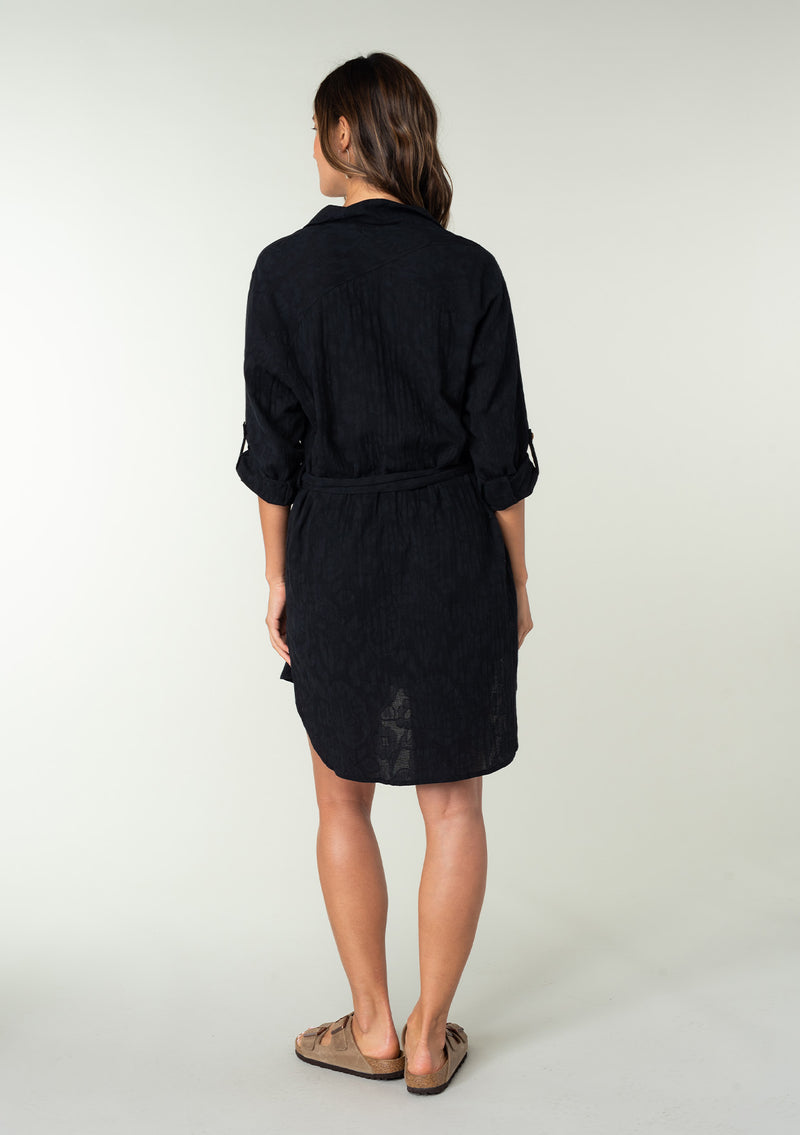 [Color: Black] A back facing image of a brunette model wearing a bohemian black cotton tunic shirt in a textured jacquard. With long rolled sleeves and a button tab closure, a collared neckline, a button front, a self tie waist belt, side vents, a round hemline, and a back yoke detail.