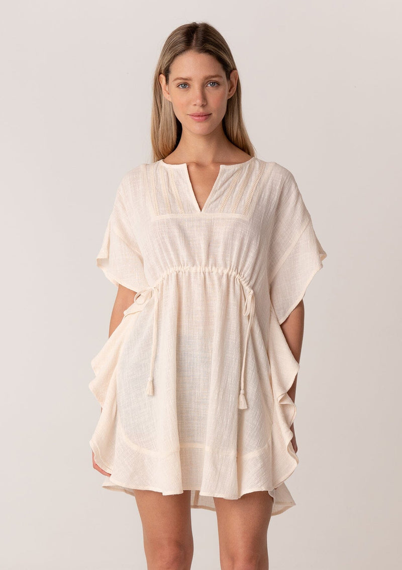 [Color: Natural] A front facing image of a blonde model wearing a bohemian caftan top in an off white cotton. A beach cover up style with short sleeves, a ruffled hemline, a v neckline, embroidered details throughout, and a drawstring waist detail in the front and back with tassel ties. 