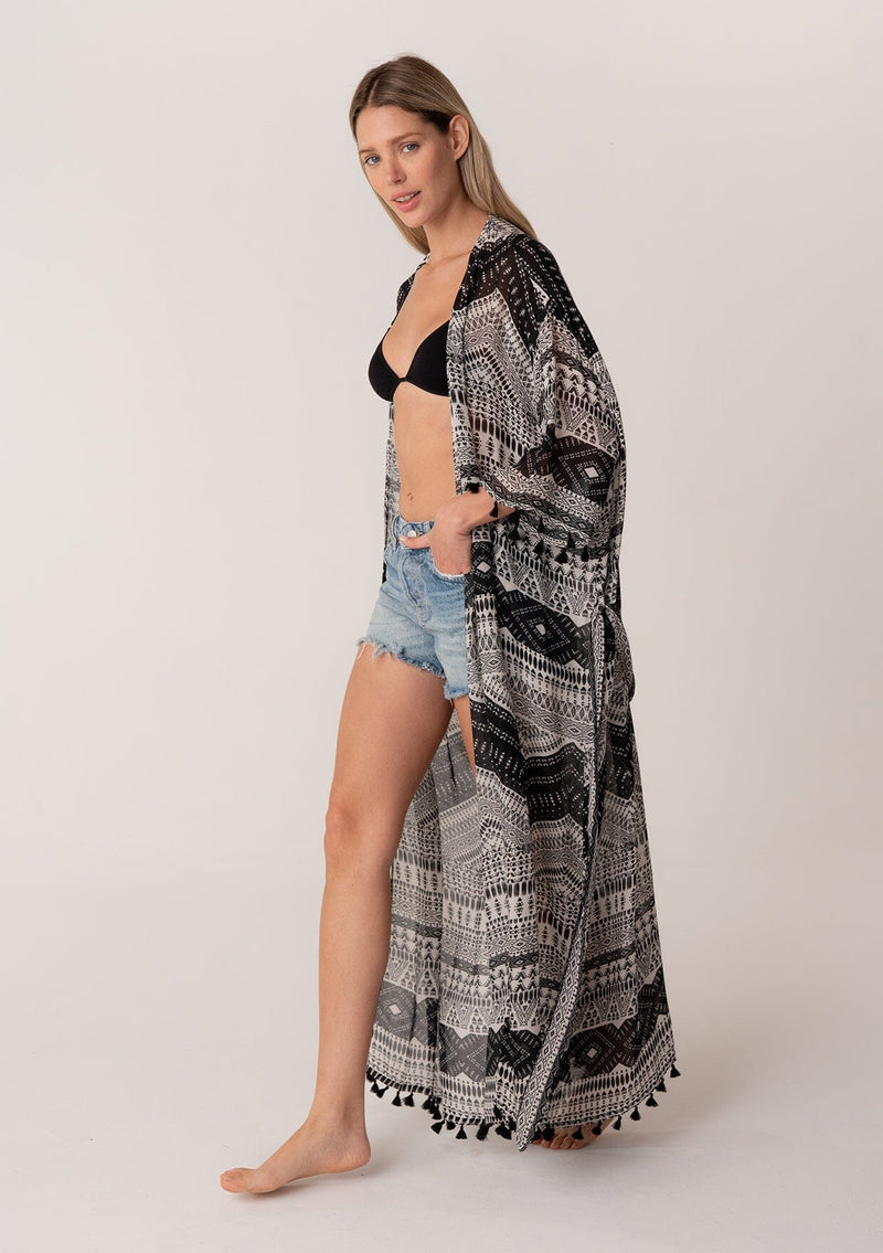 [Color: Black/Natural] A side facing image of a blonde model wearing a black printed bohemian resort kimono. A maxi length cover up with tassel fringe hem, half length sleeves, and a tie waist belt. 