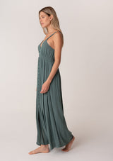 [Color: Lagoon] A side facing image of a blonde model wearing a bohemian sleeveless spring maxi dress in dusty teal. With a self covered button front, a deep v neckline, a front slit, a smocked elastic empire waist, and an open back with elastic strap detail.