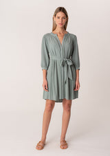 [Color: Sea Green] A full body front facing image of a blonde model wearing a dusty teal bohemian spring mini dress. With three quarter length long sleeves, lattice lace trim details, a concealed button front, and a self tie waist belt.