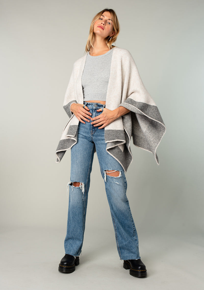 [Color: Heather Natural/Heather Grey] A full body front facing image of a blonde model wearing a soft and warm mid length sweater cape. An open front cape cardigan with a contrast border design. Perfect fall sweater, great for layering. 
