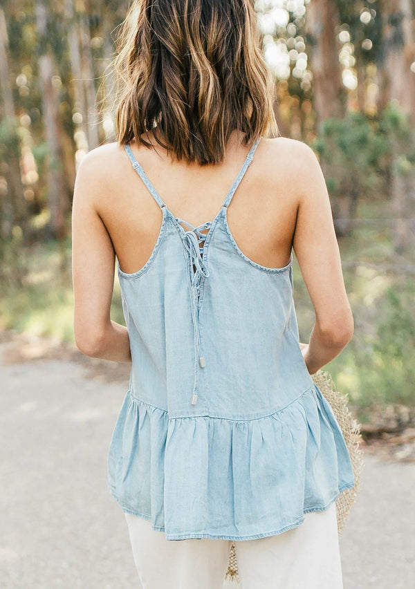 [Color: Heritage Blue] A blond woman wearing a flowy denim like tank top with a criss cross lace up back detail, adjustable spaghetti straps, and a tiered hemline.