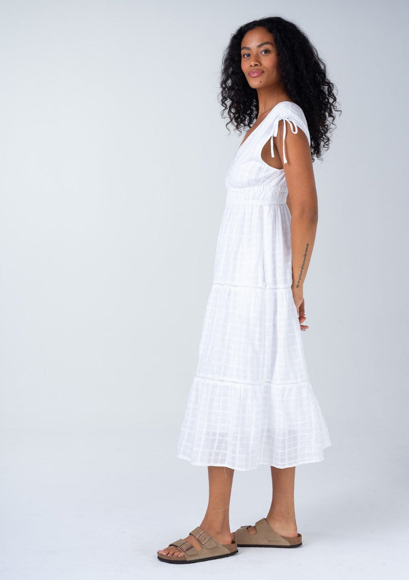 [Color: White] A side facing image of a brunette model wearing a classic bohemian spring mid length dress in a white cotton textured gingham. With adjustable shoulder ties, a v neckline in front and back, a lace trimmed tiered skirt, an elastic waist, and side pockets. 