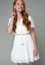 [Color: Off White] A front facing image of a red headed model wearing a white eyelet mini dress with short flutter sleeves, a split v neckline with tassel ties, and an elastic waist. The model has long red wavy hair. 