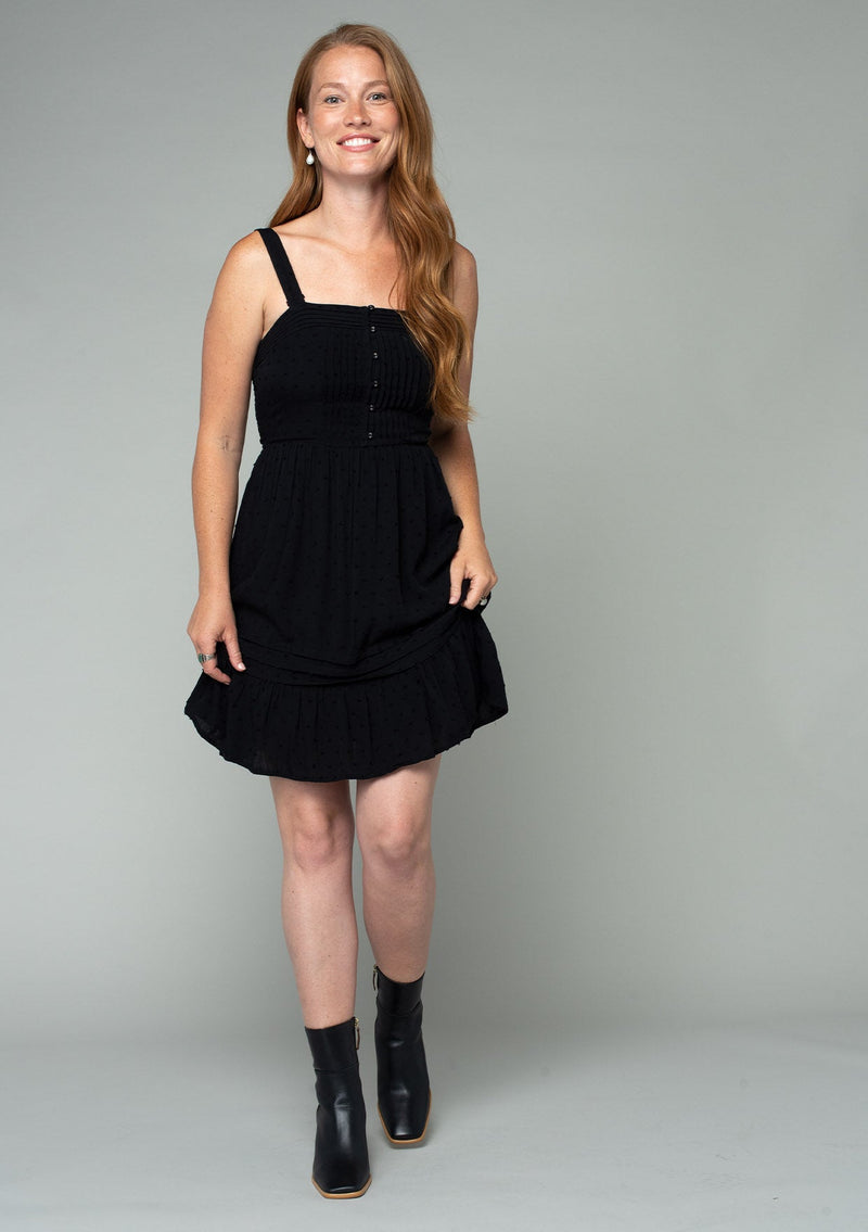 [Color: Black] A full body front facing image of a red headed model wearing a black sleeveless bohemian mini dress with a button front top, tank top straps, a square neckline, pleated pintuck details, and a flowy mini skirt.