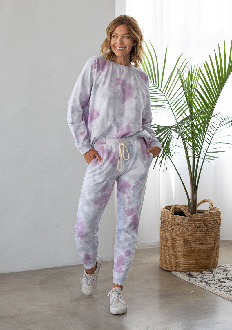 [Color: Lavender Combo] A woman standing outside wearing a classic cotton jogger pant in a splatter tie dye wash. Featuring a drawstring waistband, side pockets, and a slim leg.