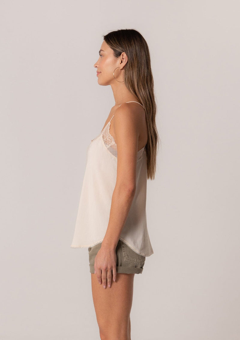 [Color: Vanilla] A side facing image of a brunette model wearing an ivory camisole tank top. With spaghetti straps, a v neckline, lace trim, a racerback, and a relaxed fit.