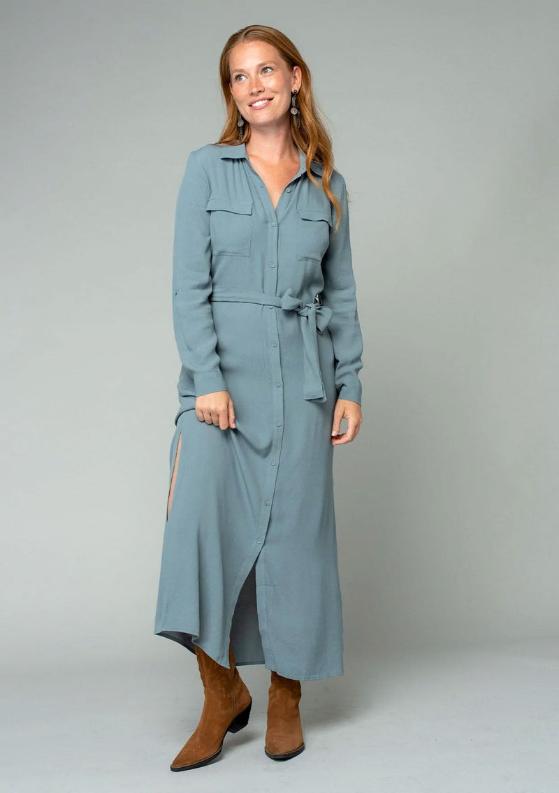 [Color: Balsam Green] A front facing image of a red headed model wearing a relaxed fit light green maxi shirt dress made from a lightweight crepe. With long rolled sleeves, a button tab sleeve closure, two front pockets, side pockets at the hip, and a self tie waist belt.