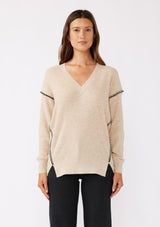 [Color: Oatmeal/Black] A brunette model wearing an everyday sweater with a relaxed silhouette in a beige tan. With a classic v neckline, long sleeves, and an exposed seam with blanket stitch. A fall sweater styled with black bottom and casual sandals. 