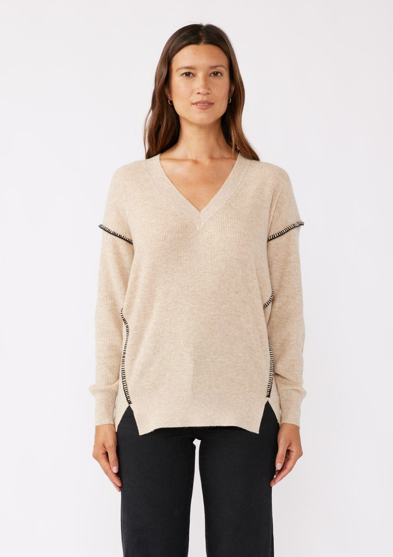 [Color: Oatmeal/Black] A brunette model wearing an everyday sweater with a relaxed silhouette in a beige tan. With a classic v neckline, long sleeves, and an exposed seam with blanket stitch. A fall sweater styled with black bottom and casual sandals. 