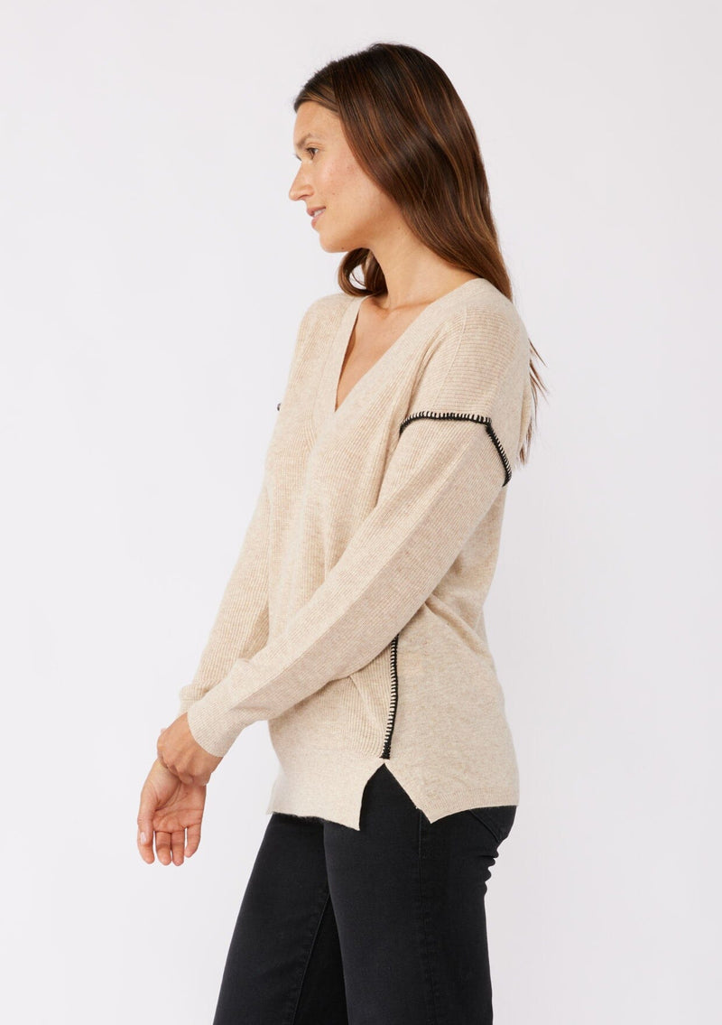 [Color: Oatmeal/Black] A brunette model wearing an everyday sweater with a relaxed silhouette in a beige tan. With a classic v neckline, long sleeves, and an exposed seam with blanket stitch. A fall sweater styled with black bottom and casual sandals. 