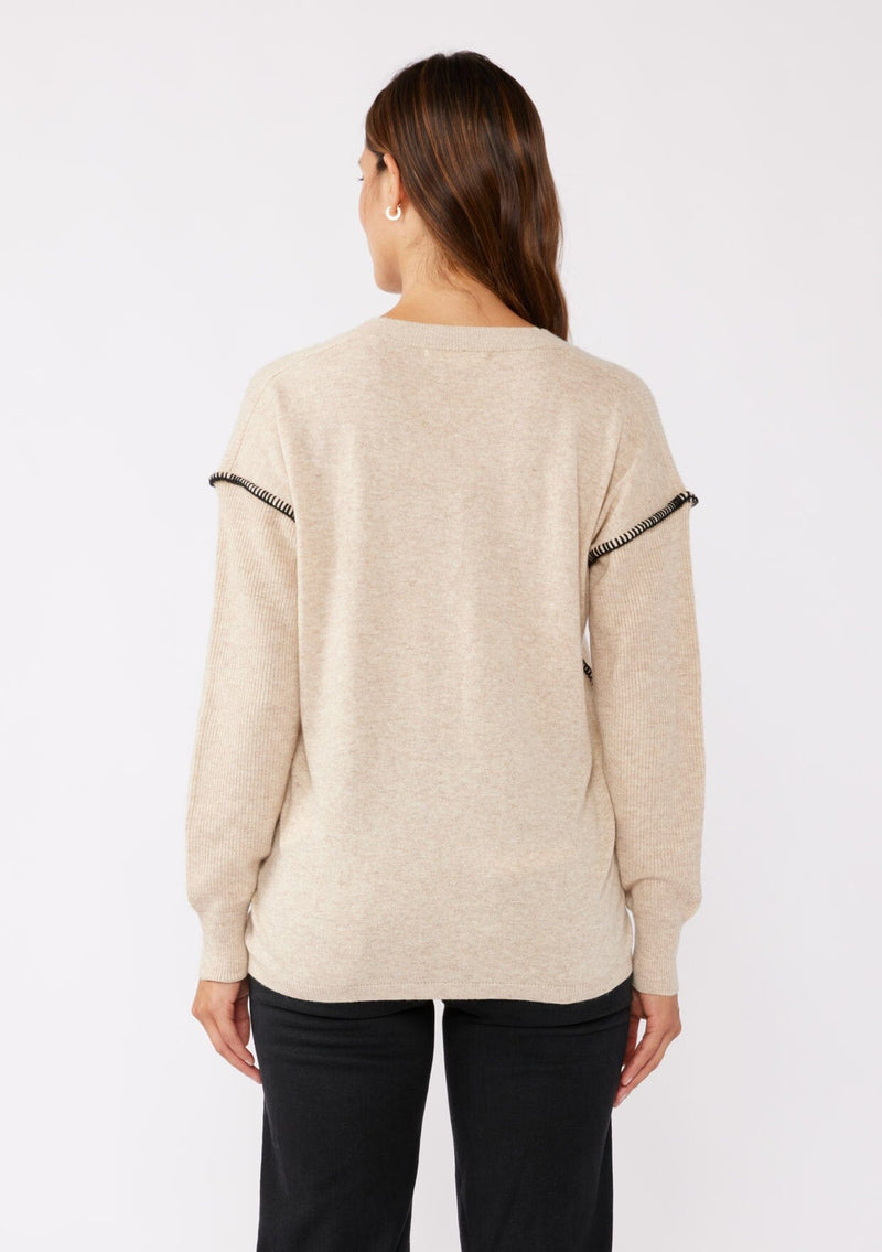 [Color: Oatmeal/Black] A brunette model wearing an everyday sweater with a relaxed silhouette in a beige tan. With a classic v neckline, long sleeves, and an exposed seam with blanket stitch. A fall sweater styled with black bottom and casual sandals. 