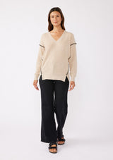[Color: Oatmeal/Black] A brunette model wearing an everyday sweater with a relaxed silhouette in a beige tan. With a classic v neckline, long sleeves, and an exposed seam with blanket stitch. A fall sweater styled with black bottom and casual sandals. 