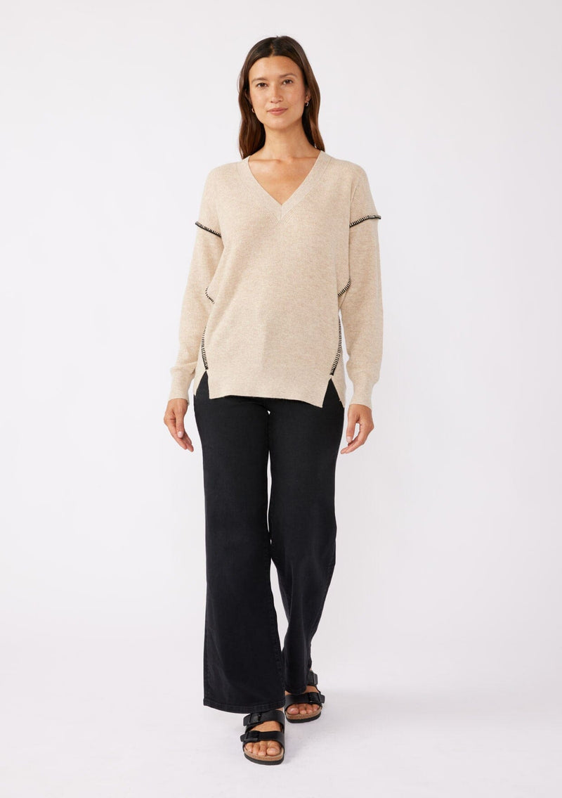 [Color: Oatmeal/Black] A brunette model wearing an everyday sweater with a relaxed silhouette in a beige tan. With a classic v neckline, long sleeves, and an exposed seam with blanket stitch. A fall sweater styled with black bottom and casual sandals. 
