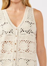 [Color: Natural] A detail image of a blonde model wearing a button-front vest crafted in a cotton floral crochet pattern. Featuring a sleeveless design and flattering V-neckline, this breezy resort vest is perfect for beach days and vacation styling.
