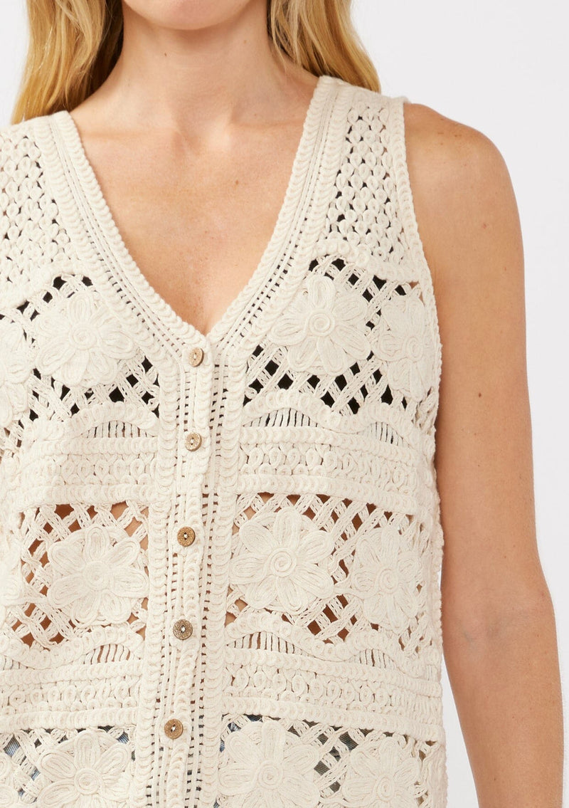 [Color: Natural] A detail image of a blonde model wearing a button-front vest crafted in a cotton floral crochet pattern. Featuring a sleeveless design and flattering V-neckline, this breezy resort vest is perfect for beach days and vacation styling.