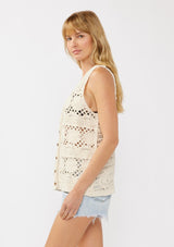 [Color: Natural] A side facing image of a blonde model wearing a button-front vest crafted in a cotton floral crochet pattern. Featuring a sleeveless design and flattering V-neckline, this breezy resort vest is perfect for beach days and vacation styling.