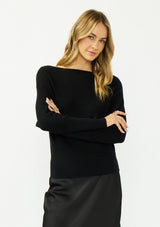 [Color: Black] A front facing image of a blonde model wearing a black pullover sweater with a boat neckline and long sleeves that taper at the cuffs. Styled with a black satin skirt. 